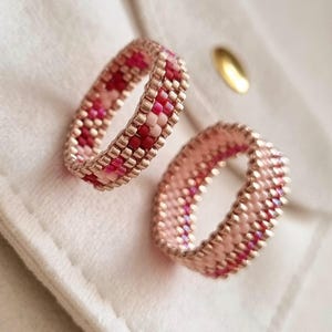 May include: Two beaded rings crafted from small, uniform glass beads. One ring features a geometric pattern of alternating pink, light pink, and deep red beads. The second ring displays a solid light pink band with a thin, vertical stripe of red beads.