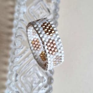 May include: A beaded ring with a repeating pattern of white, gold, and rose gold beads. The ring is set against a blurred background of a clear glass object.