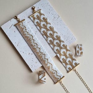 Può includere: Un set di gioielli con due braccialetti di perline, un anello e una piccola perla rettangolare. I braccialetti e l'anello hanno un motivo bianco e oro. I braccialetti hanno chiusure e prolunghe a catena dorate. La piccola perla rettangolare ha un motivo simile.