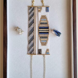 May include: Two beaded bracelets and two beaded rings displayed in a wooden frame. The bracelets feature intricate patterns in shades of gray, blue, and gold. The rings have similar designs. The jewelry is set against a white background.