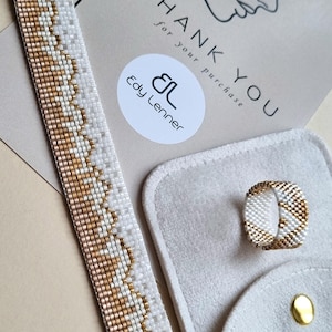 May include: A beaded bracelet and ring set in white and gold, with a geometric pattern. The jewellery is displayed with a thank you card and a small grey pouch. The bracelet is approximately 2.5 cm wide.