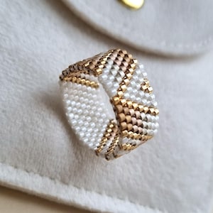 May include: A beaded ring with a geometric pattern. The ring features alternating rows of white, gold, and tan beads. The ring is set against a white background.