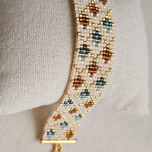 May include: A woven bead bracelet featuring a cream-colored background with a repeating geometric pattern of metallic gold, copper, and teal beads. The bracelet is finished with a gold-toned metal clasp and a lobster claw closure.