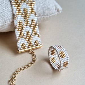 Könnte beinhalten: Ein Armband und Ring-Set aus Perlen. Das Armband ist weiß mit einem goldenen geometrischen Muster und einem goldenen Verschluss. Der Ring hat ein passendes Design. Der Schmuck wird auf neutralem Hintergrund präsentiert.