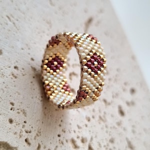 May include: A beaded ring featuring a repeating pattern of burgundy and gold beads. The ring is primarily cream-colored with gold accents and a textured surface in the background.