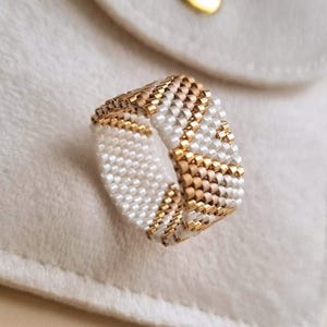 May include: A wide band ring crafted from small, cylindrical beads arranged in a geometric pattern. The design features alternating sections of matte white beads and metallic gold beads, creating a textured, woven appearance across the entire surface.