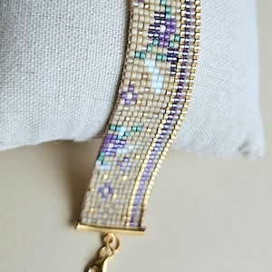 May include: Close-up of a beaded bracelet with a gold clasp. The bracelet features a geometric pattern in cream, purple, teal, and gold. The beads are small and tightly woven together, creating a textured surface. The clasp is a simple hook and eye design.