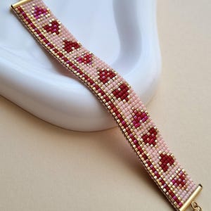 May include: A beaded bracelet with a pink and red heart design. The bracelet is made of small, colourful beads and has gold-coloured metal accents and a clasp. The bracelet is approximately 2 cm wide.