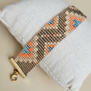 Puede incluir: Una pulsera de cuentas con un patrón geométrico en tonos beige, marrón, naranja y azul. La pulsera tiene un cierre dorado y se muestra sobre una superficie blanca texturizada. La pulsera mide aproximadamente 2 cm de ancho.