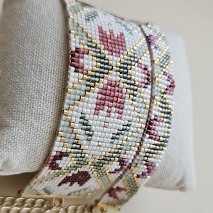 May include: A beaded bracelet with a floral pattern in white, pink, green, and gold. The bracelet is displayed on a beige pillow and has a gold chain clasp. The beads are small and tightly woven.