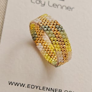 May include: A beaded ring featuring a geometric pattern of metallic gold, pale yellow, white, and muted sage green seed beads. The ring is displayed on a white card with the text Edy Lenner and the website URL WWW.EDYLENNER.COM printed in black.