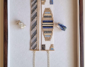 May include: Two rectangular beaded bracelets and two matching beaded rings. One bracelet features a diagonal gray and gold pattern, while the other has a central geometric design in blue, gold, and black. Both bracelets have gold-tone metal ends and extender chains.