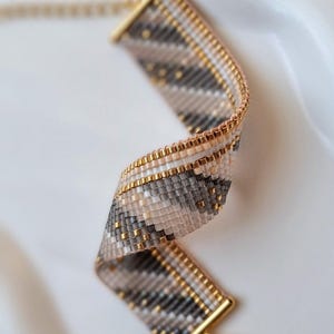 Op de afbeelding: Een geweven kralenarmband met een diagonaal streeppatroon in grijstinten, beige en wit, geaccentueerd met metallic gouden kralen. De armband is afgewerkt met goudkleurige metalen eindstukken en een bijpassende karabijnsluiting.