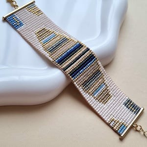 Może przedstawiać: Tkana bransoletka z geometrycznym wzorem. Bransoletka ma jasnoróżową bazę ze złotymi, niebieskimi i czarnymi koralikami. Posiada złote metalowe końcówki i zapięcie na łańcuszek.