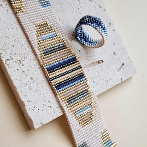 Puede incluir: Una pulsera de cuentas y un anillo a juego con un patrón geométrico de cuentas de vidrio en color crema, dorado y varios tonos de azul. La pulsera tiene terminales de metal dorado y un cierre de mosquetón, el anillo es una banda continua.