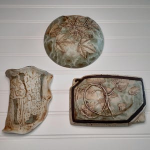 May include: Three ceramic wall art pieces with leaf and vine designs. The pieces are in shades of green, brown, and cream. One is round with a leaf pattern, another is rectangular with vines, and the third is an irregular shape with text.