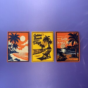 May include: Three rectangular embroidered patches featuring beach scenes. The left patch shows palm trees and a sun in orange and navy. The centre patch is yellow with palm trees, a chair, and the text Golden Beach Days. The right patch depicts a sunset with the text Salt In the Air.
