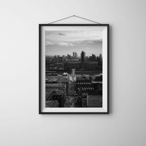 Può includere: Stampa incorniciata in bianco e nero di un paesaggio urbano. L'immagine presenta un fiume con un ponte e degli edifici sullo sfondo. La stampa è esposta su una parete bianca.
