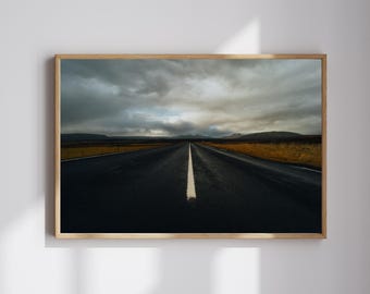 May include: A framed photograph of a road disappearing into the distance under a dramatic, cloudy sky. The dark asphalt road has a white central line. The landscape includes fields and mountains, creating a sense of depth and vastness.