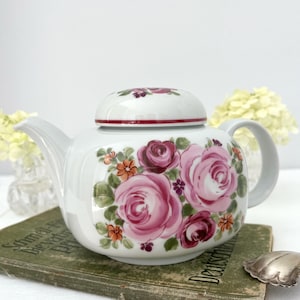 Könnte beinhalten: Eine weiße Porzellanteekanne mit einem handgemalten Blumenmuster aus großen rosa und dunkelroten Rosen, kleinen orangefarbenen Blüten und grünen Blättern. Die Teekanne hat einen passenden Deckel mit einem dünnen roten Rand.