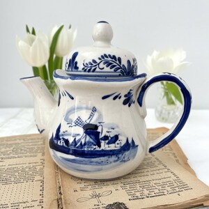 Peut inclure: Une théière en céramique blanche avec des motifs floraux et de moulins à vent bleus. La théière a une anse incurvée et un couvercle avec un petit bouton. Le design rappelle la poterie de Delft.