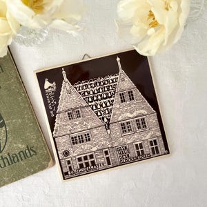 Könnte beinhalten: Quadratische, schwarz-weiße Fliese mit einer detaillierten Illustration eines Gebäudes mit mehreren Fenstern und einem Satteldach. Die Fliese trägt deutschen Text, darunter "Das Haus des Glockenspiels" und "Böttcherstrasse". Es ist ein dekoratives Stück.