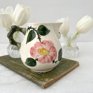 Könnte beinhalten: Cremefarbener Keramikkrug mit einem rosa Blumen- und grünen Blättermuster. Der Krug hat einen dunkelgrünen Griff und steht auf einem alten Buch. Weiße Tulpen in kleinen Glasvasen sind im Hintergrund.