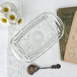Pode incluir: Uma manteigueira retangular de vidro transparente com um padrão decorativo. Uma colher de prata vintage com uma tigela em forma de concha e detalhes florais está por perto. Margaridas num pequeno vaso de vidro e um livro vintage também estão presentes.