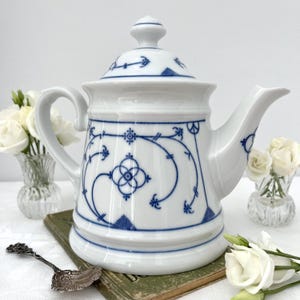 May include: A white porcelain teapot featuring a blue floral and geometric pattern, a matching lid with a knob handle, a curved spout, and a side handle. The teapot is decorated with intricate blue line work and symmetrical botanical motifs.