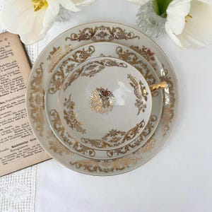 Könnte beinhalten: Ein cremefarbenes Vintage-Teeservice mit goldenen Blumen- und Scrollarbeiten. Das Set besteht aus einer Tasse, einer Untertasse und einem Teller. Die Tasse hat einen goldenen Henkel. Das Set ist auf einer weißen Oberfläche angeordnet.