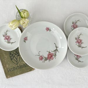 Op de afbeelding: Een verzameling witte porseleinen borden en schotels met een roze bloemenontwerp. De set bevat een groot dinerbord, verschillende kleinere borden en schotels. Een vintage boek en een kleine vaas met bloemen zijn ook aanwezig.