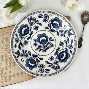 Op de afbeelding: Witte keramische plaat met een blauw bloemmotief. De plaat heeft een centraal bloemmotief en een rand van kleine blauwe bloemen. Het ontwerp doet denken aan vintage servies.