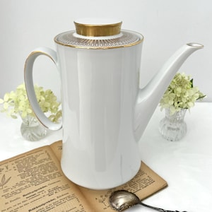 May include: A white porcelain coffee pot featuring a tall cylindrical body, a curved handle, and a long spout. The lid is accented with a gold rim and a decorative band of geometric patterns in gold and black. The pot has a smooth, glossy finish.