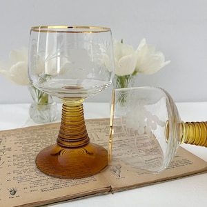 Könnte beinhalten: Zwei klare Weingläser mit geätzten Blumendesigns und goldenen Rändern. Die Stiele sind bernsteinfarben mit gerippter Textur. Ein Glas steht aufrecht, das andere liegt auf der Seite. Die Gläser stehen auf einem Vintage-Buch mit weißen Blumen im Hintergrund.