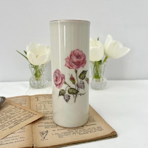 Può includere: Un vaso cilindrico color crema con bordo dorato, ornato da illustrazioni di rose rosa. Il vaso è alto circa 20 cm e poggia su un libro aperto con illustrazioni botaniche. Tulipani bianchi in piccoli vasi di vetro sullo sfondo.