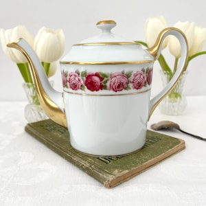 May include: White porcelain teapot with a gold handle and spout, adorned with a band of pink and red roses. The lid and rim are trimmed with gold. The teapot sits on a vintage green book, with white tulips in the background.