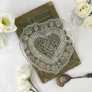 Könnte beinhalten: Eine klare, herzförmige Glasschale mit gepresstem Rauten- und Gittermuster, die auf einem alten grünen Buch mit dem Titel Schmeil-Fitschen liegt. Die Schale hat einen gewellten Rand und ist von weißen Rosen sowie einem silberfarbenen Metalllöffel umgeben.