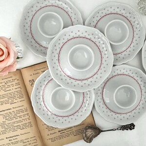 May include: A set of four white ceramic egg cups with scalloped edges, featuring a delicate pink dotted pattern and a central circular indentation. Each piece has a smooth, glossy finish and a consistent design, arranged in a cluster.