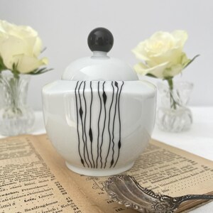 Può includere: Una zuccheriera in ceramica bianca con un pomello nero sul coperchio. La ciotola presenta un design a linee nere verticali. La ciotola è su un libro d'epoca con un cucchiaio d'argento.