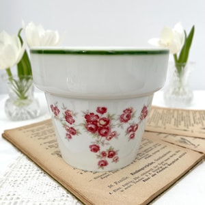 Può includere: Un vaso di fiori in ceramica bianca con bordo verde e motivo floreale di rose rosse. Il vaso è su un libro d'epoca. Il vaso è progettato per piantare fiori. Misure in centimetri.