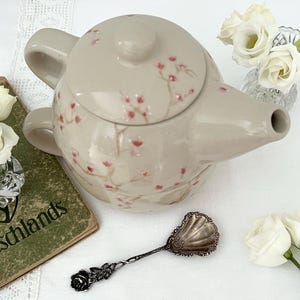 Può includere: Una teiera in ceramica color beige chiaro con un delicato motivo dipinto a mano di piccoli fiori di ciliegio rosa sul coperchio e sul corpo. La teiera presenta un manico curvo e un beccuccio, accompagnata da un cucchiaio in metallo argentato.