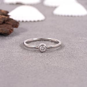 May include: A silver ring featuring a small, round, clear gemstone at its centre. The ring's band is slender and polished. The ring is presented on a grey surface, with white seashells and a piece of wood in the background.