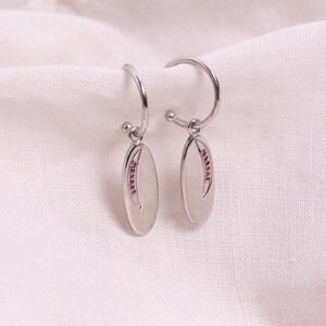 May include: A pair of silver hoop earrings with oval pendants. Each pendant has a line of small, pink gemstones. The earrings are set against a white, textured fabric background.