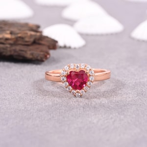 May include: A rose gold ring with a heart-shaped, bright red gemstone, encircled by a halo of smaller, clear gemstones. The ring is set on a light grey surface, with a blurred background.