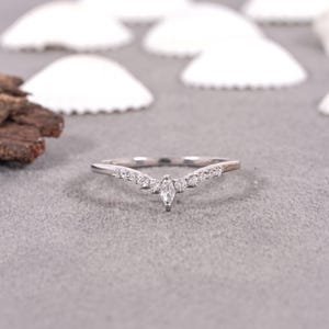 Puede incluir: Un anillo plateado con un diseño curvo, engastado con múltiples piedras preciosas pequeñas y transparentes. El anillo presenta una piedra central en forma de marquesa y se exhibe sobre una superficie gris con acentos de conchas blancas.