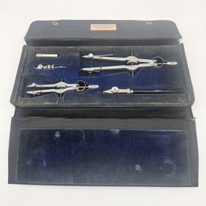 May include: A vintage drafting instrument set housed in a black case with a blue velvet interior. The set includes two silver metal compasses, a small metal attachment, and a black pen-like tool with the text VEMCO printed on the side.