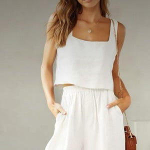 May include: White linen crop top and wide-leg pants set. The top has a square neckline. The pants have an elastic waistband and pockets. A small brown purse hangs from a shoulder strap.