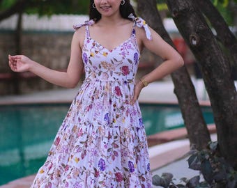 May include: A white floral print maxi dress with a V-neckline and tie straps. The dress features a tiered skirt and is adorned with purple, pink, and yellow floral patterns. The dress is a summer style.