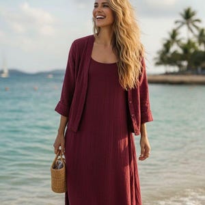 May include: A burgundy dress and cardigan set is worn by a woman on a beach. The dress is midi length with a scoop neck. She holds a small woven basket. The background shows the sea and palm trees.