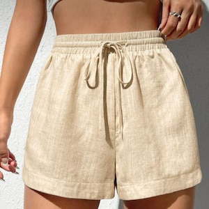 Puede incluir: Pantalones cortos de lino beige con cintura elástica y cordón. Los pantalones cortos tienen un ajuste relajado y bolsillos laterales. La tela tiene una textura sutil y los pantalones cortos parecen ligeros y adecuados para el clima cálido.